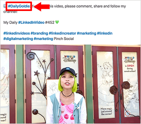 Dit is een screenshot dat illustreert hoe Goldie Chan hashtags gebruikt in de tekst van haar LinkedIn-videoposts. Rode callouts verwijzen naar de hashtag #DailyGoldie in de tekst, die uniek is voor haar videoposts en haar helpt bij het bijhouden van shares. Het bericht bevat ook andere relevante hashtags waarmee mensen haar video kunnen vinden, waaronder #LinkedInVideo. Op het videobeeld staat Goldie voor enkele deuren bij een World of Disney-display. Ze is een Aziatische vrouw met groen haar. Ze draagt ​​een zwarte pet van LinkedIn, een zwarte halsketting, een roze overhemd met macaronprint en een blauw-wit jasje.